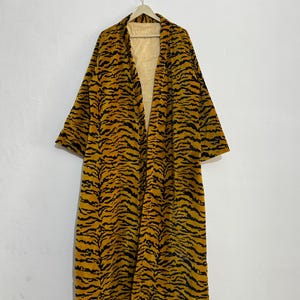 May include: A long, open-front robe with a tiger stripe pattern in shades of yellow and black. The robe is hanging on a wooden hanger against a white wall. The interior lining is a light yellow with vertical stripes.