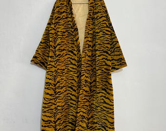 May include: A long, open-front robe with a tiger stripe pattern in shades of yellow and black. The robe is hanging on a wooden hanger against a white wall. The interior lining is a light yellow with vertical stripes.