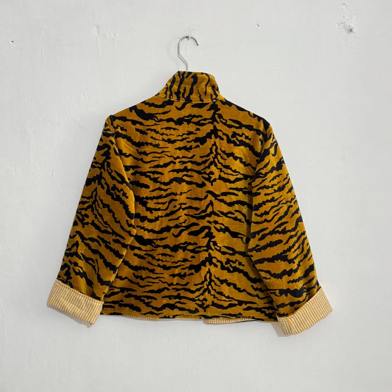 May include: A mustard yellow jacket with a black tiger stripe pattern. The jacket has a high collar and long sleeves with striped cuffs. The jacket is hanging on a metal hanger against a white wall.