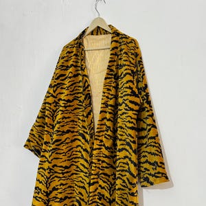 May include: A long, open-front robe with a tiger stripe pattern in shades of yellow and black. The robe is hanging on a wooden hanger against a white wall. The interior lining is a light yellow with vertical stripes.
