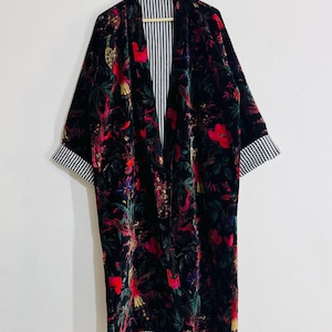 May include: A long, black velvet robe with a vibrant floral pattern in red, green, and yellow. The robe features a striped lining and cuffs. The robe is hanging on a wooden hanger against a white background.