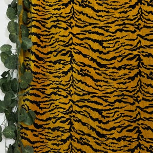 Puede incluir: Primer plano de una tela con un estampado de rayas de tigre. La tela tiene un fondo amarillo con rayas negras. Hay vides verdes y frondosas en el lado izquierdo de la imagen.