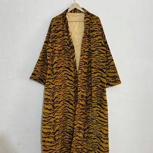 May include: A long, open-front robe with a tiger stripe pattern in black and gold. The robe is hanging on a wooden hanger against a white wall. The interior lining is a light yellow striped fabric.