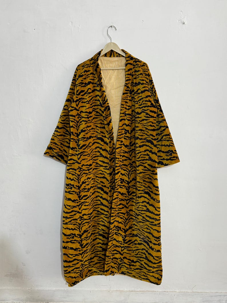 May include: A long, open-front robe with a tiger stripe pattern in black and gold. The robe is hanging on a wooden hanger against a white wall. The interior lining is a light yellow striped fabric.