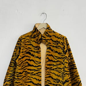 May include: A reversible jacket with a tiger stripe pattern. The outer layer is a mustard yellow with black stripes, and the inner layer is a light yellow with vertical stripes. The jacket is hanging on a metal hanger against a white wall.