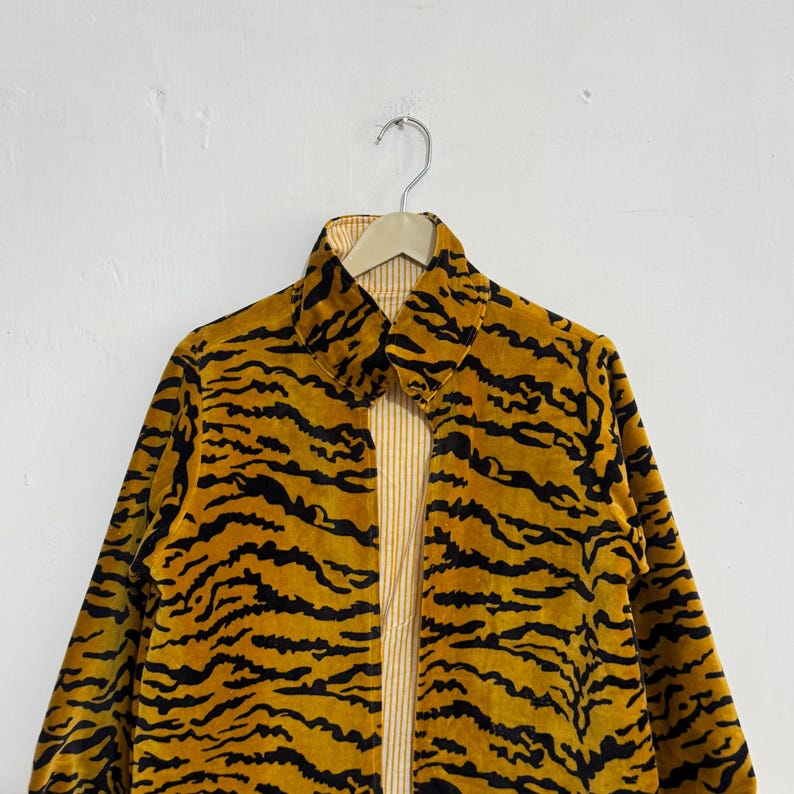 May include: A reversible jacket with a tiger stripe pattern. The outer layer is a mustard yellow with black stripes, and the inner layer is a light yellow with vertical stripes. The jacket is hanging on a metal hanger against a white wall.