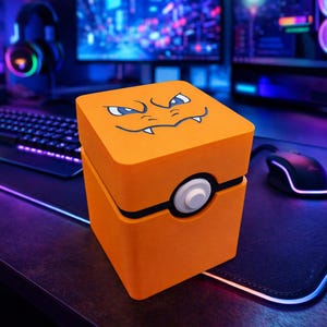 May include: An orange cube-shaped storage box featuring a cartoon face on the lid. The box has a white and black circular design on the front. The box is on a desk with a keyboard, mouse, and headphones in the background.