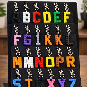 May include: A display of colorful Lego-style letter keychains. Each letter is a different color and attached to a silver keychain. The text at the top says "LEGO STYLE LETTER KEYCHAINS" and "Pick your letter & color!"