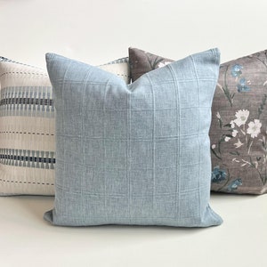 May include: Four decorative throw pillows in various patterns and colors. The front pillow is a light blue with a grid pattern. Other pillows feature a white and blue geometric design, a floral pattern, and a brown and white floral pattern.