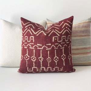 May include: A burgundy throw pillow with a white geometric pattern. The pillow is surrounded by two other pillows, one white and one striped.