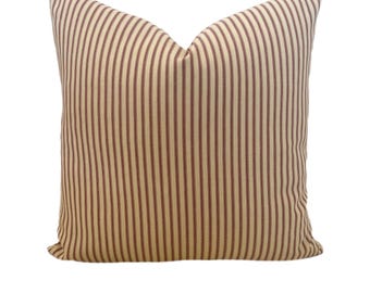 May include: A square pillow with a cream-colored background and vertical burgundy stripes. The pillow has a soft, textured appearance and is designed for decorative use. The pillow is a neutral color and would complement a variety of home decor styles.