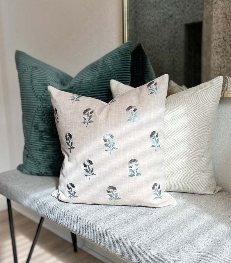K&ouml;nnte beinhalten: Drei dekorative Kissen auf einer grauen Bank. Das mittlere Kissen ist beige mit einem floralen Muster in Schwarz und Blau. Das Kissen links ist aus dunkelgr&uuml;nem Cord. Das Kissen rechts hat ein wei&szlig;-graues Fischgr&auml;tenmuster.