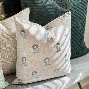 K&ouml;nnte beinhalten: Drei dekorative Kissen auf einer grauen und wei&szlig;en gestreiften Bank. Das Kissen in der Mitte ist ein hellbeiges Leinenkissen mit einem floralen Muster in Grau- und Blaut&ouml;nen. Das Kissen links ist ein wei&szlig;-graues Fischgratmuster. Das Kissen rechts ist ein dunkelgr&uuml;nes Cordkissen.