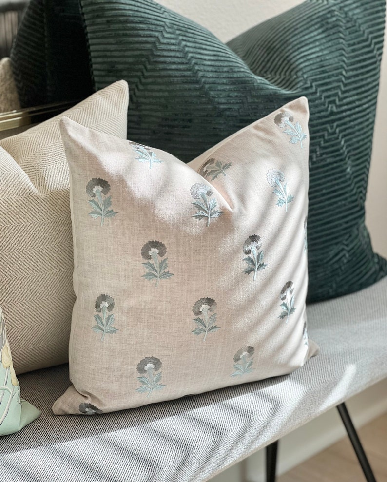 K&ouml;nnte beinhalten: Drei dekorative Kissen auf einer grauen und wei&szlig;en gestreiften Bank. Das Kissen in der Mitte ist ein hellbeiges Leinenkissen mit einem floralen Muster in Grau- und Blaut&ouml;nen. Das Kissen links ist ein wei&szlig;-graues Fischgratmuster. Das Kissen rechts ist ein dunkelgr&uuml;nes Cordkissen.