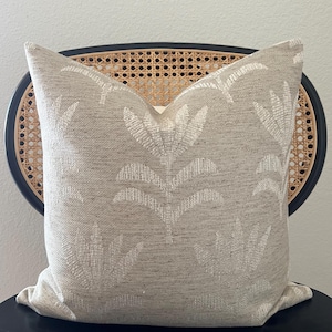 May include: A beige decorative pillow with a white floral pattern. The pillow is square and sits on a black chair with a woven back. The pillow's fabric has a textured appearance, and the floral design is repeated across the surface.