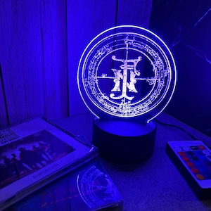 Könnte beinhalten: Blaue LED-Lampe mit kreisförmigem Design und kompliziertem Symbol. Die Lampe steht auf einem schwarzen Sockel, neben einer Fernbedienung und einer CD-Hülle. Der Raum wird von blauem Licht beleuchtet.