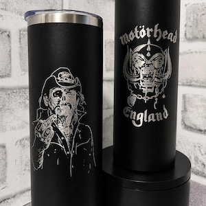 May include: Two matte black cylindrical tumblers with silver lids and straws. The left tumbler features a detailed engraving of a bearded figure in a hat. The right tumbler displays the Motörhead logo with a skull graphic and the text England.