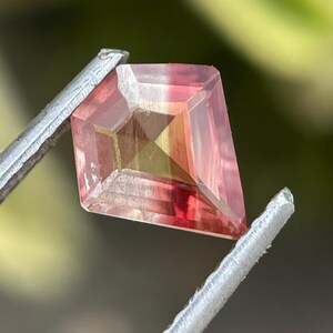 May include: A kite-shaped gemstone featuring a gradient of colors transitioning from a soft yellow at the center to a deep pink at the edges. The stone has a faceted surface that reflects light, creating distinct geometric patterns across its face.
