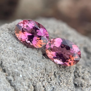 Puede incluir: Dos gemas de corte ovalado con un degradado de tonos rosa y naranja. Las gemas se encuentran sobre una superficie de piedra gris rugosa, resaltando sus facetas pulidas y sus colores vibrantes. Las gemas parecen ser turmalinas.