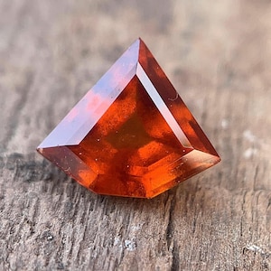 Könnte beinhalten: Ein facettierter, drachenförmiger Edelstein mit einem warmen, rötlich-orangen Farbton. Die scharfen Winkel und polierten Oberflächen des Edelsteins reflektieren das Licht und erzeugen ein brillantes Aussehen. Der Edelstein liegt auf einer strukturierten Holzoberfläche.
