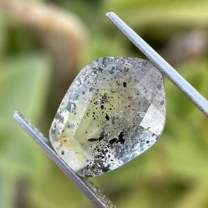 May include: A faceted, translucent gemstone with a pale yellow tint, containing numerous small, dark, irregular inclusions scattered throughout its interior. The stone is held by a pair of silver-toned metal tweezers.