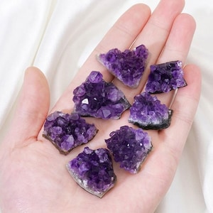 Op de afbeelding: Een verzameling ruwe amethistkristallen in verschillende vormen en maten. De kristallen vertonen een diepe paarse tint met enkele gebieden in lichter violet. De kristallen worden in een hand gehouden, wat hun natuurlijke, ongepolijste staat laat zien. De kristallen hebben een ruw, getextureerd oppervlak.