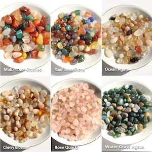 May include: Six white bowls filled with polished stones. The stones are labeled: Multi-color Crushed, Rainflower Stone, Ocean Agate, Cherry Blossom, Rose Quartz, and Water Grass Agate. The stones vary in color, including red, green, pink, and white.