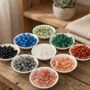 May include: A collection of small, round, colorful beads in various shades, including black, blue, green, white, gray, orange, and red, are displayed in small white bowls. The beads are arranged on a wooden surface, suggesting a craft or jewelry-making theme.