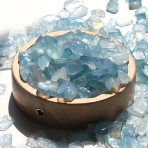 May include: A wooden bowl filled with light blue tumbled gemstones. The gemstones are scattered around the bowl on a white surface. The gemstones have a translucent quality and vary in size and shape.