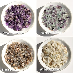 May include: Four small white plates each hold a different type of tumbled stones. The stones are Dream Amethyst, Rainbow Fluorite, Black Bone, and White Sunstone. Each plate has a gold rim and the names of the stones are written below them.
