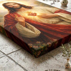 Peut inclure: Une impression sur toile religieuse repr&eacute;sentant J&eacute;sus avec un c&oelig;ur rayonnant, v&ecirc;tu d'une robe blanche et d'un manteau rouge. L'&oelig;uvre d'art est peinte en d&eacute;tail avec un &eacute;clairage doux et des motifs floraux sur le c&ocirc;t&eacute;. Un chapelet et un petit vase sont en arri&egrave;re-plan.