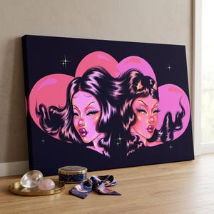 May include: A canvas art print featuring two stylized female figures with long dark hair and pink skin, set against a dark blue background with a pink heart shape. The artwork has a glamorous, illustrative style.