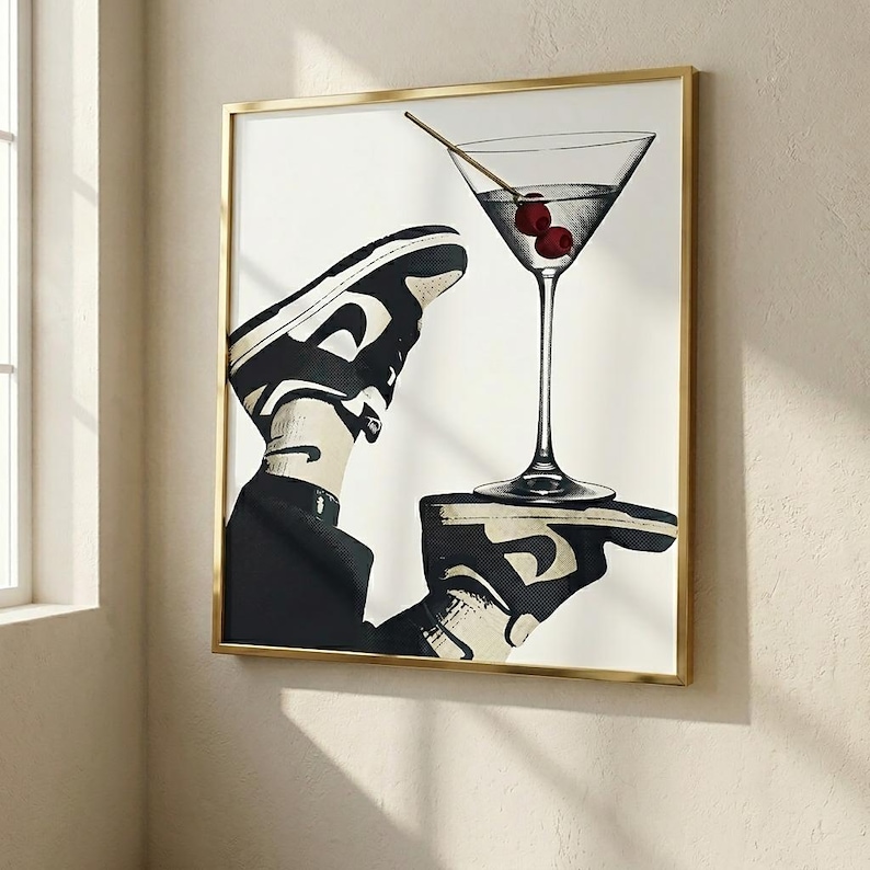 May include: A framed wall art print featuring a black and white graphic illustration of a sneaker-clad foot balancing a martini glass with two red cherries on a flat palm. The artwork is encased in a thin, metallic gold-colored rectangular frame.