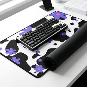 May include: A rectangular desk mat featuring a white background with black cow-print spots and purple flowers with pink centers. A black mechanical keyboard with white lettering sits on top of the mat, which is partially rolled on the right side.