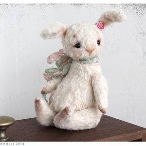 Puede incluir: Un conejito de peluche blanco con una flor rosa en la oreja y una cinta verde y rosa alrededor del cuello. El conejito est&aacute; sentado sobre una superficie de madera.