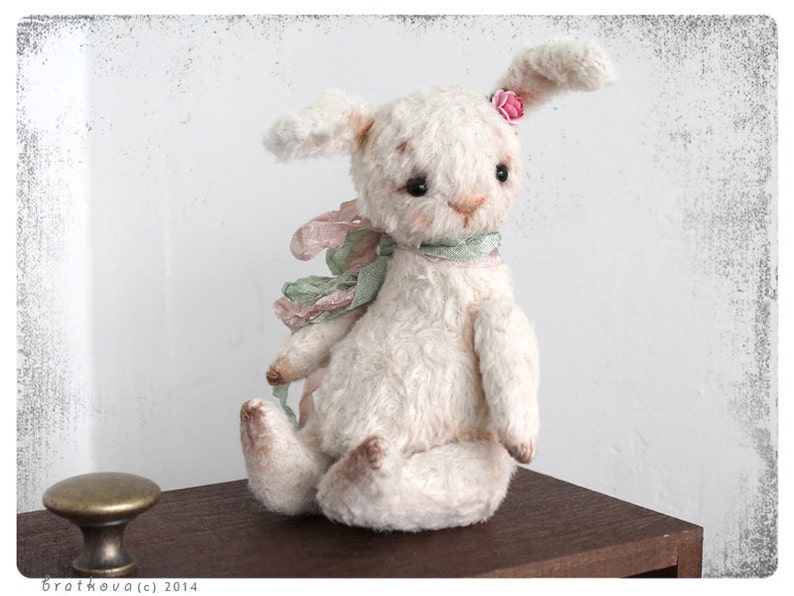 Puede incluir: Un conejito de peluche blanco con una flor rosa en la oreja y una cinta verde y rosa alrededor del cuello. El conejito est&aacute; sentado sobre una superficie de madera.