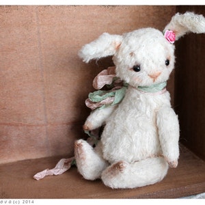 Puede incluir: Un conejito de peluche blanco con una flor rosa en la oreja, que lleva una cinta verde y rosa alrededor del cuello. El conejito est&aacute; sentado en una caja de madera.
