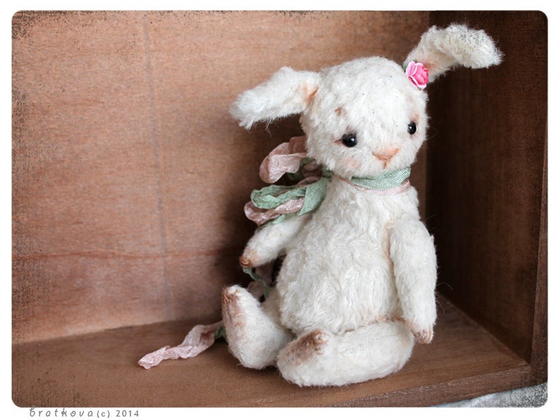 Puede incluir: Un conejito de peluche blanco con una flor rosa en la oreja, que lleva una cinta verde y rosa alrededor del cuello. El conejito est&aacute; sentado en una caja de madera.