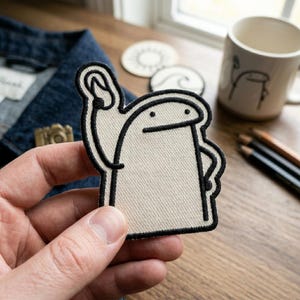 May include: A beige patch with a black outline depicting a cartoon figure waving. The patch is held in a hand, with a denim jacket and a mug in the background. Other items include coasters and pencils.