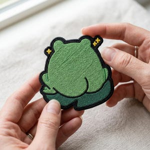 May include: A green embroidered frog patch with a black border. The frog is depicted from the back, with its legs and body visible. The patch has yellow accents and is held by two hands.