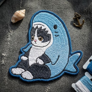 May include: Embroidered patch featuring a black and white cat wearing a blue shark costume. The patch has a light blue border and is set against a gray denim background. The cat has large, expressive eyes.