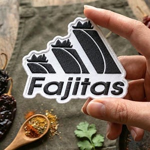 May include: A white and black patch with the word "Fajitas" in a bold font. The patch features a stylized design of a mountain range with black and white stripes. The patch is held by a hand, with spices and herbs in the background.