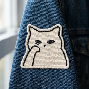 May include: A white cat patch with black outline and details is sewn onto a blue denim jacket. The cat has a slightly annoyed expression, with one paw raised. The patch is a fun, quirky accessory.
