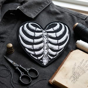 May include: Heart-shaped black patch with white rib cage embroidery. The patch is on a dark gray fabric, with a thimble, scissors, thread spools, and a sketch of a hand. The patch is a unique accessory.