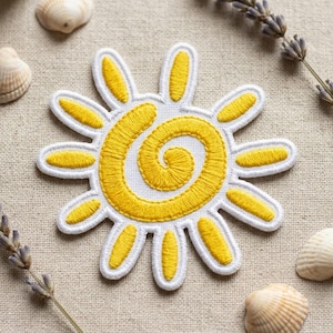 May include: Embroidered sun patch with a white border and yellow details. The sun design features a yellow spiral center and yellow rays. The patch is on a beige fabric background, with seashells and lavender sprigs.