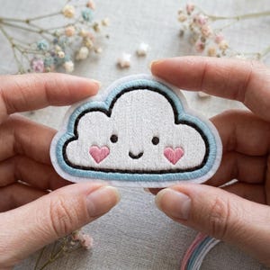 May include: A white cloud-shaped patch with a smiling face and two pink heart cheeks. The patch has a light blue border and black outline. The patch is held between two hands.