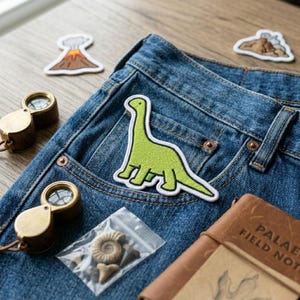May include: A light green dinosaur patch with a white border is on a blue denim pocket. Two volcano patches and two brass magnifying glasses are also visible. A small bag of fossils and a brown leather notebook are also in the image.