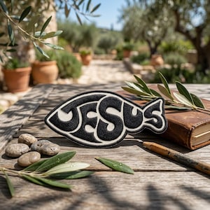 May include: Black and white embroidered patch in the shape of a fish, with the word "JESUS" in a stylized font. The patch is on a wooden surface with a book, pencil, and olive branches.