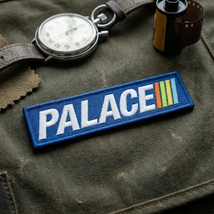 May include: A blue embroidered patch with the word "PALACE" in white block letters. The patch has a rectangular shape with a colorful striped design. A stopwatch and film canister are in the background.