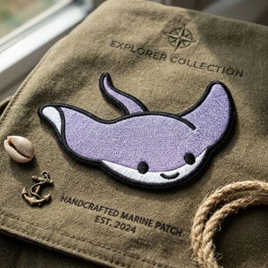 May include: A purple and white embroidered manta ray patch with a black outline, placed on a green canvas. The text "EXPLORER COLLECTION" and "HANDCRAFTED MARINE PATCH EST. 2024" are visible. A seashell and anchor charm are also present.